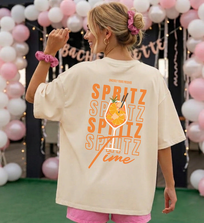 Beiges T-Shirt mit großem, orangefarbenem Spritz Time Rückendruck und Cocktail-Grafik, präsentiert in einer festlichen Dekoration mit Luftballons.
