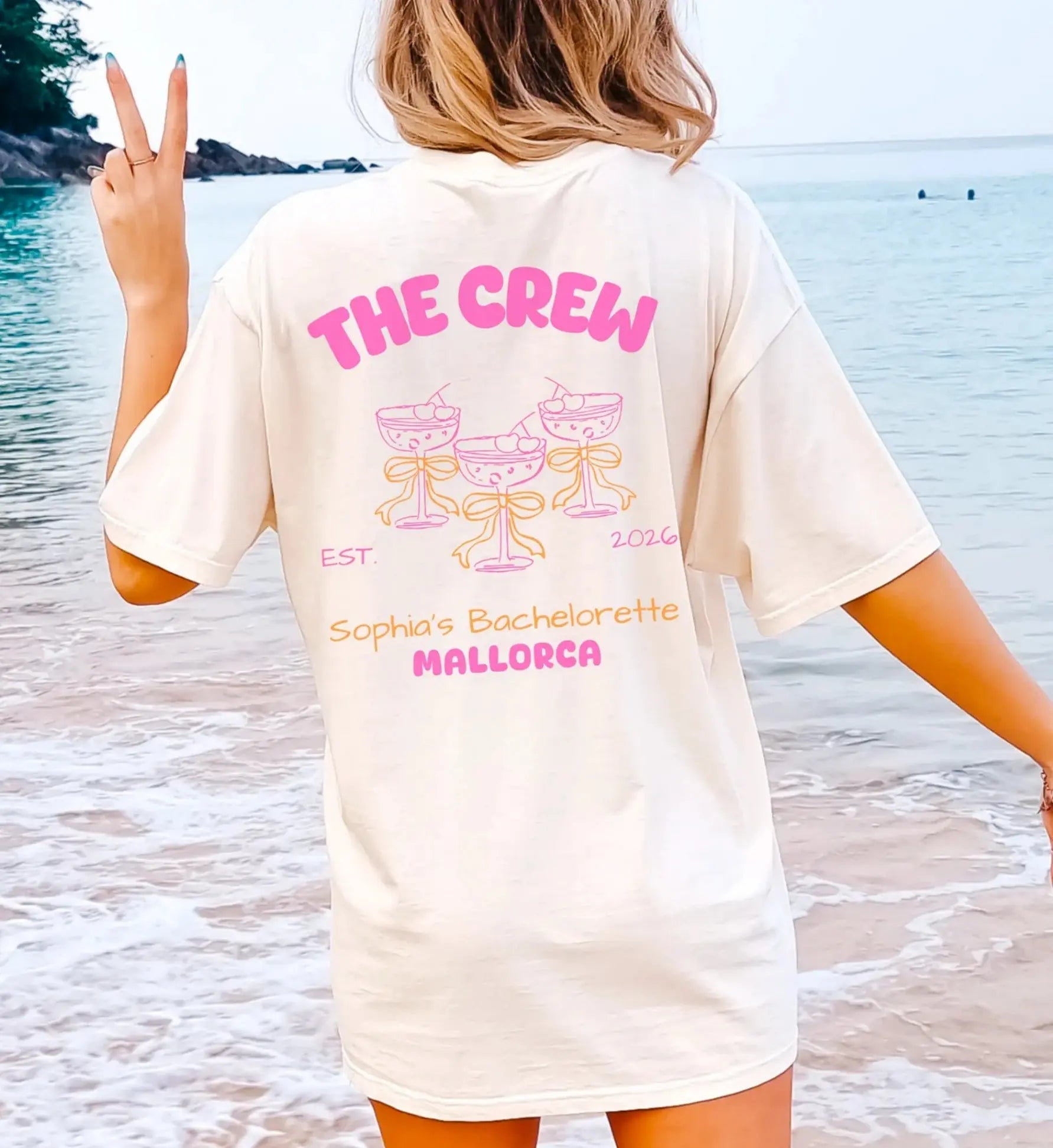 Cremefarbenes JGA T-Shirt für Damen mit 'The Crew' Rückenprint, illustrierten Cocktailgläsern mit gelben Schleifen im Coquette-Stil, getragen am Strand.