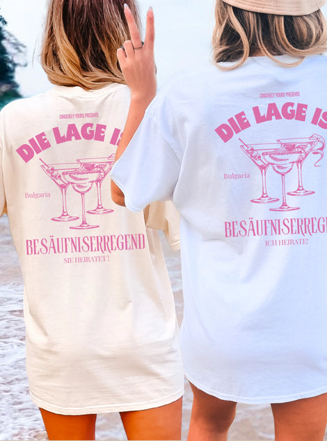 Zwei Frauen am Strand in personalisierten JGA Shirts von Cincerely Yours im Old Money Stil, Rückenansicht mit pinkem Cocktail-Aufdruck und dem Spruch 'Die Lage ist besäufniserregend' für den Junggesellinnenabschied.