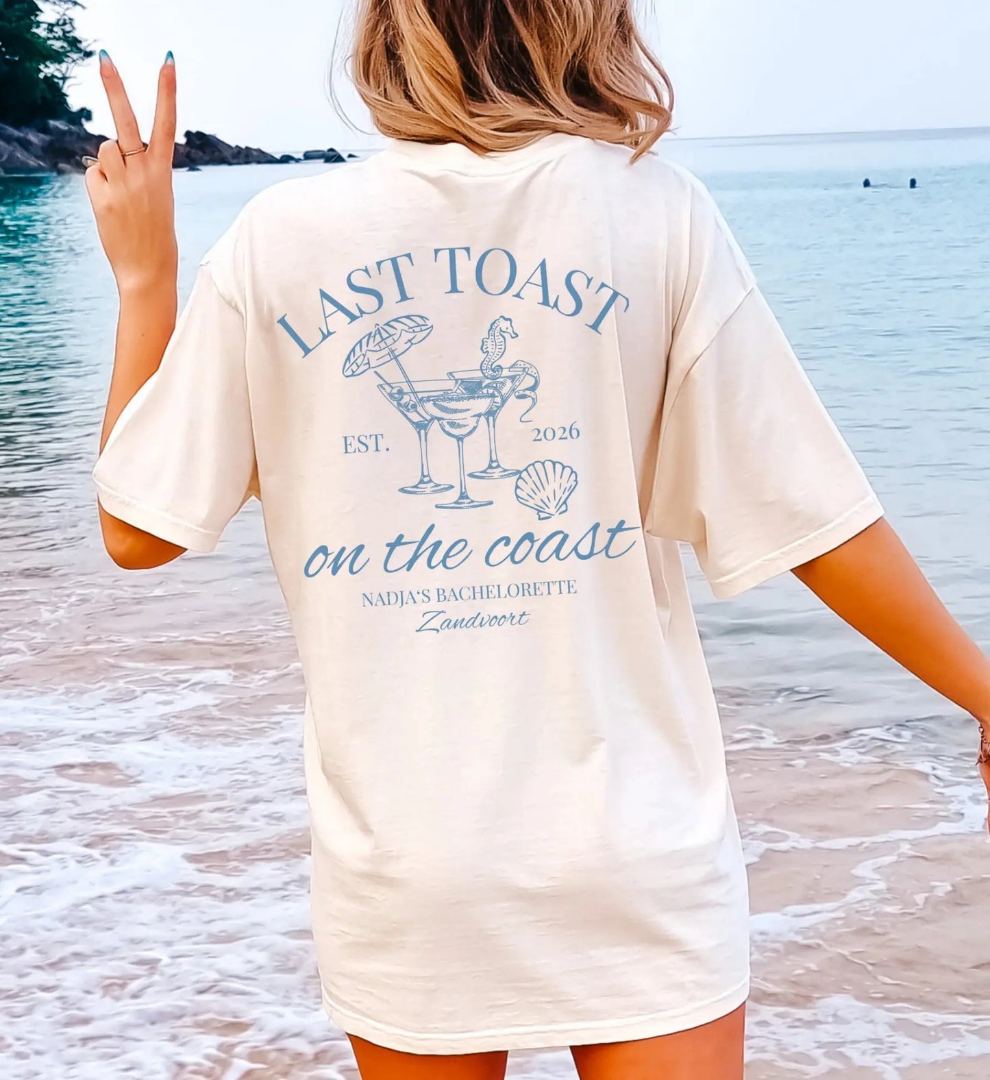 Rückansicht eines cremefarbenen Oversize T-Shirts mit hellblauem 'Last Toast on the Coast' Aufdruck, Cocktails und maritimen Motiven am Strand für einen Junggesellinnenabschied.