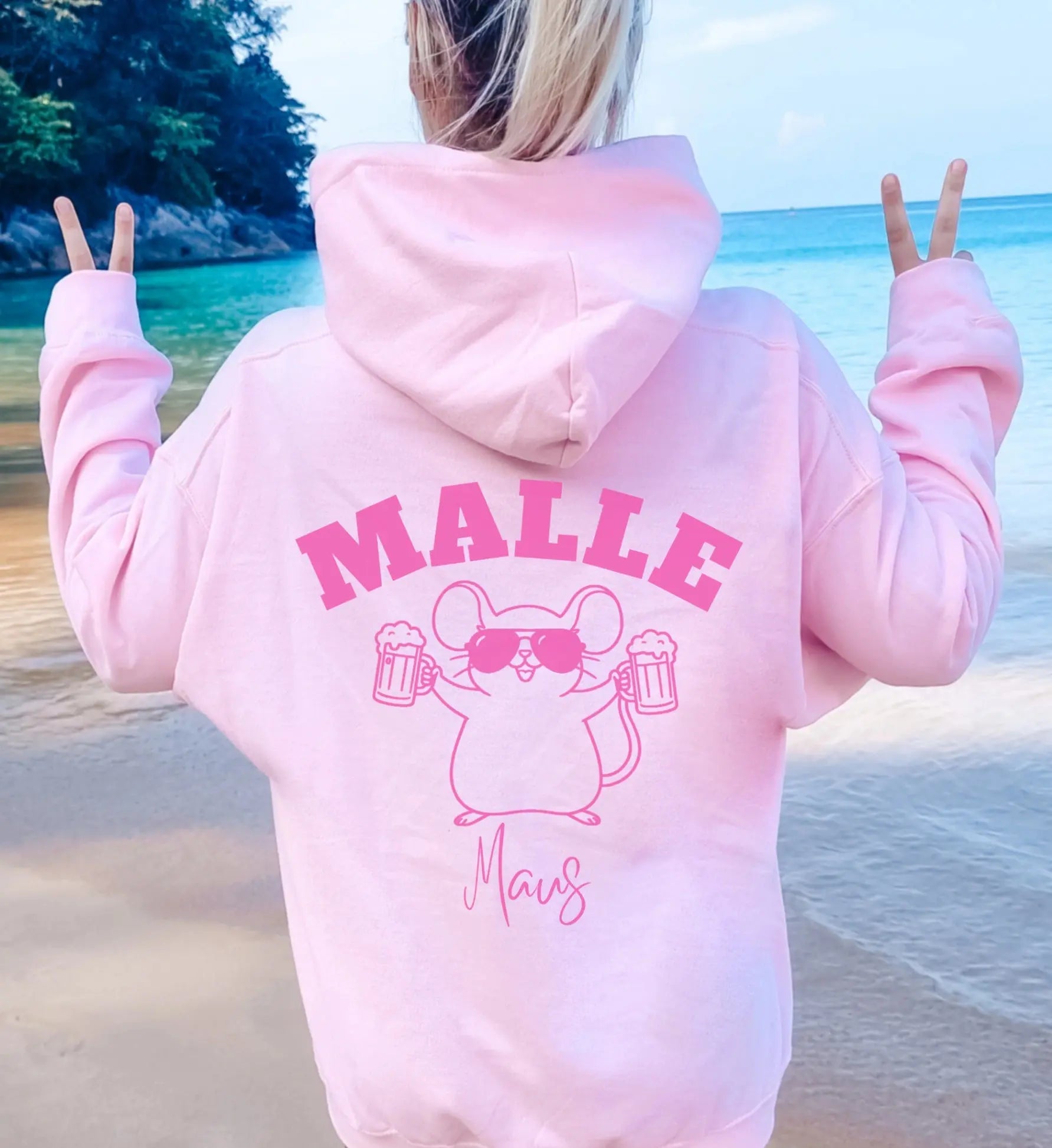 Hellrosa Oversized Hoodie mit Malle Maus Print auf der Rückseite, getragen von einer Frau am Strand mit Blick auf das türkisfarbene Meer.