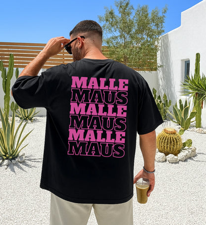 Mann in der Rückansicht trägt ein schwarzes Oversized T-Shirt mit pinkem 'Malle Maus' Grafikprint in einer mediterranen Gartenanlage.