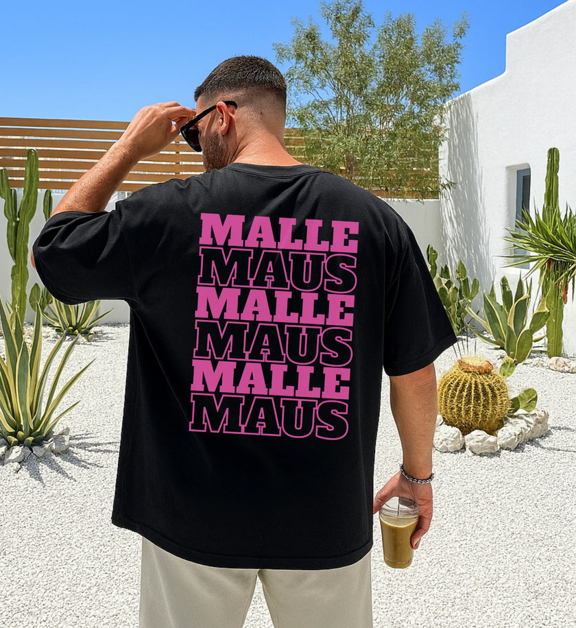 Mann in der Rückansicht trägt ein schwarzes Oversized T-Shirt mit pinkem 'Malle Maus' Grafikprint in einer mediterranen Gartenanlage.