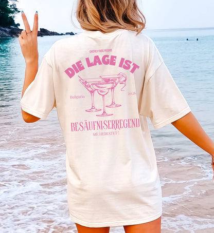 Personalisiertes JGA Shirt von Cincerely Yours im Old Money Stil mit pinkem Cocktail-Print und dem Spruch 'Die Lage ist besäufniserregend - Sie heiratet', Frau am Strand in der Rückenansicht.