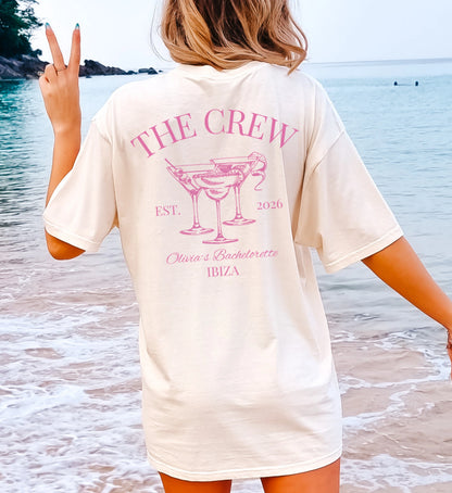 Personalisierter JGA Shirt Backprint im Old Money Stil: Cremefarbenes T-Shirt mit pinkem 'The Crew' Aufdruck, Cocktail-Gläsern und Ibiza-Schriftzug, getragen von einer Frau am Strand.