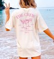 Personalisierter JGA Shirt Backprint im Old Money Stil: Cremefarbenes T-Shirt mit pinkem 'The Crew' Aufdruck, Cocktail-Gläsern und Ibiza-Schriftzug, getragen von einer Frau am Strand.