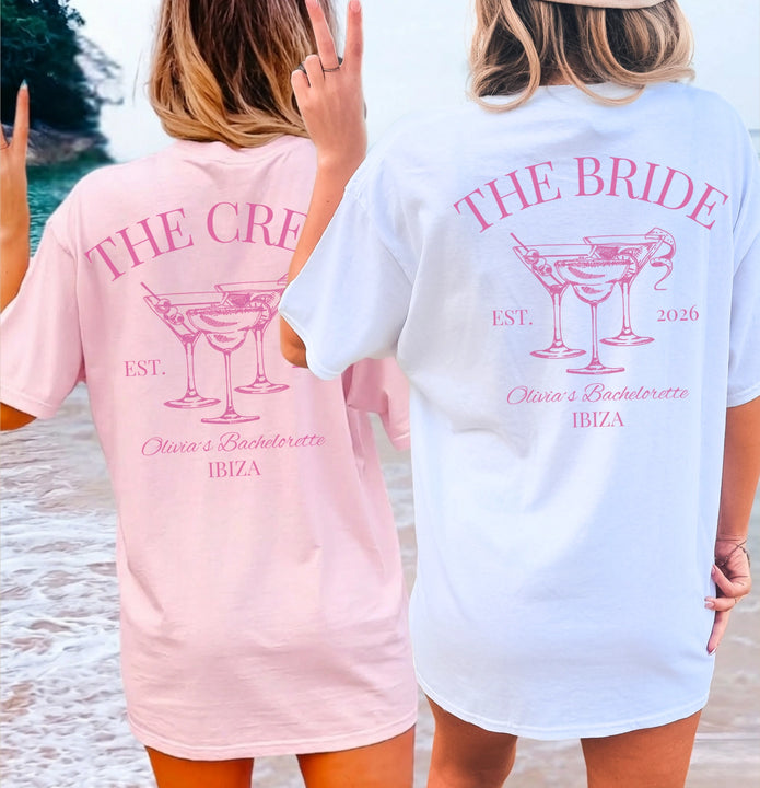 Zwei Frauen am Strand tragen personalisierte Old Money Bachelorette Shirts von Cincerely Yours. Die T-Shirts in Weiß und Rosa zeigen einen Backprint mit 'The Bride' und 'The Crew' sowie Martini-Gläsern für einen Junggesellinnenabschied in Ibiza.