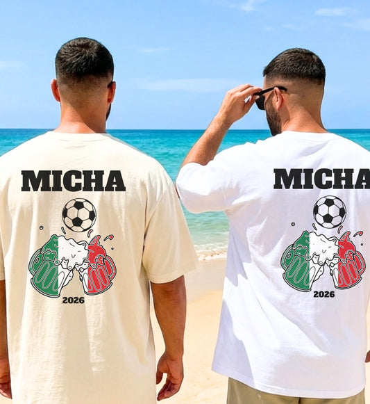 Zwei Männer am Strand tragen personalisierte T-Shirts mit einem Grafikdesign aus zwei anstoßenden Bierkrügen in den Farben der italienischen Flagge, einem Fußball und der Jahreszahl 2026.