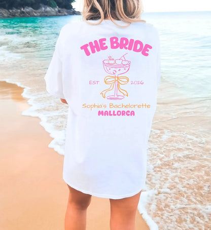Oversized weißes JGA T-Shirt für die Braut mit pinkem Mallorca-Aufdruck, Cocktail-Grafik und Schleife am Strand.
