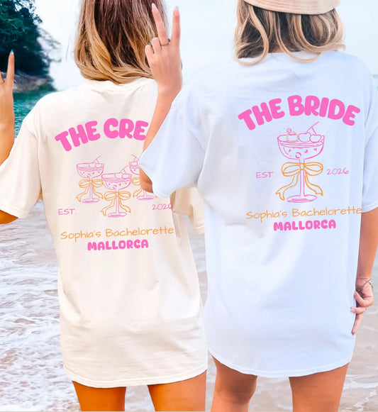 Zwei Frauen am Strand in personalisierten JGA T-Shirts für Braut und Crew mit modernem Cocktail- und Schleifen-Design im Coquette-Stil.