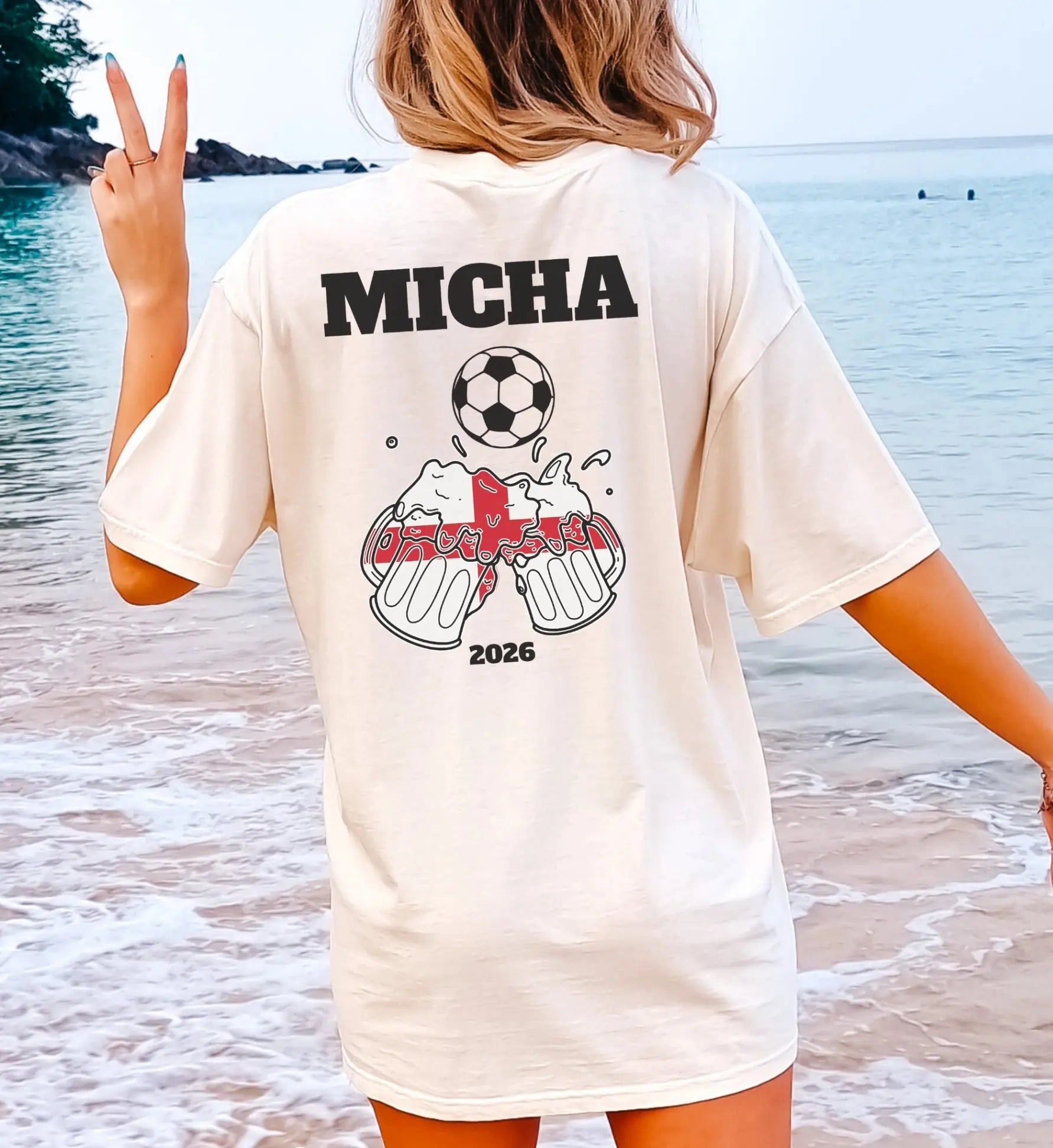 Person am Strand trägt ein personalisiertes creme-farbenes England Fußball Fan Shirt mit dem Namen Micha, einem Fußball-Motiv und anstoßenden Bierkrügen in Form der englischen Flagge.