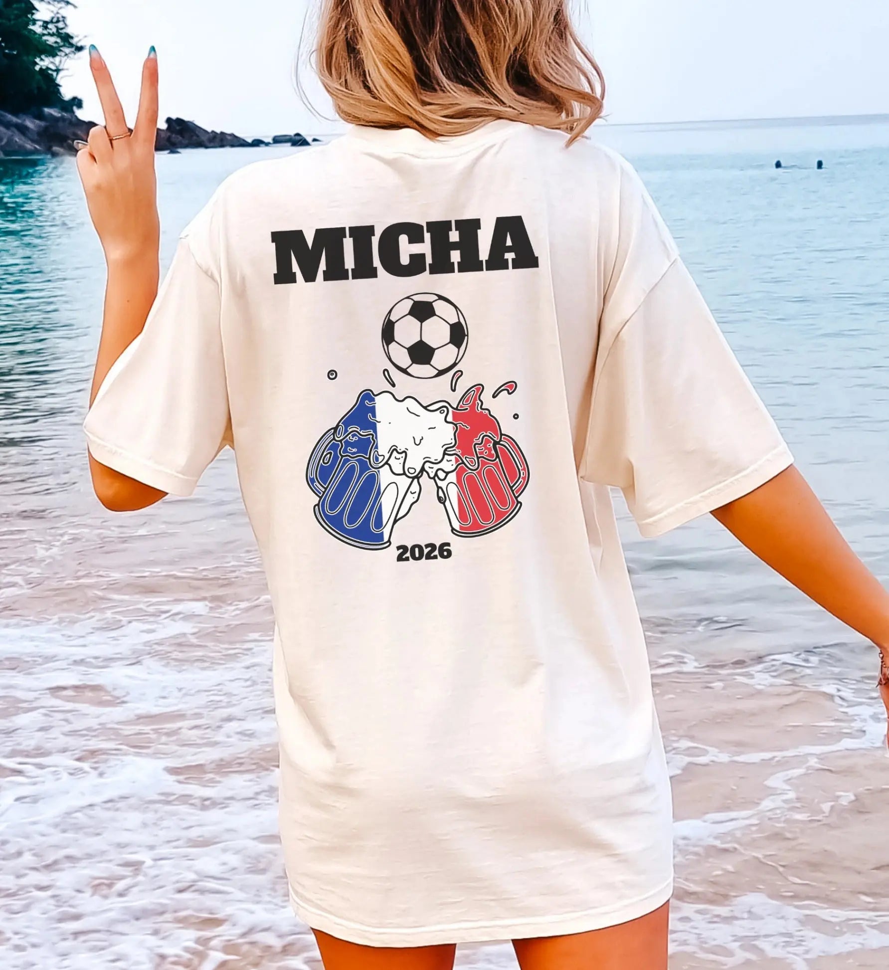 Personalisierbares weißes T-Shirt mit dem Namen 'MICHA', einem Fußball und zwei anstoßenden Bierkrügen in den französischen Nationalfarben vor einer sonnigen Strandkulisse.