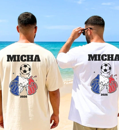 Zwei Männer am Strand in personalisierten T-Shirts für Fußballfans mit dem Namen Micha, einem Fußball-Motiv, anstoßenden Bierkrügen in den Farben Blau-Weiß-Rot und der Jahreszahl 2026.