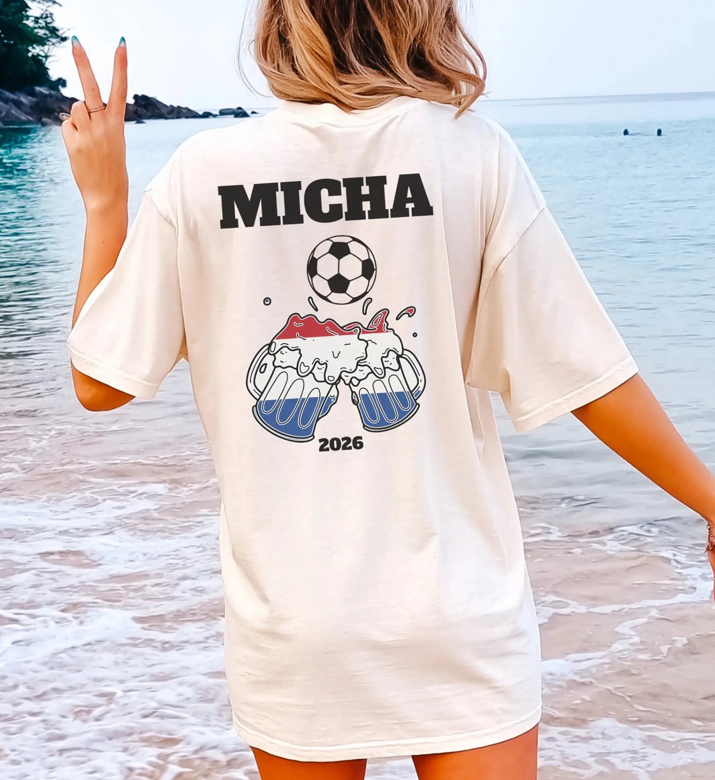 Person am Strand trägt ein personalisiertes, cremefarbenes Holland Fußball T-Shirt mit dem Namen Micha, einem Fußball-Motiv und anstoßenden Bierkrügen in den niederländischen Nationalfarben für das Jahr 2026.