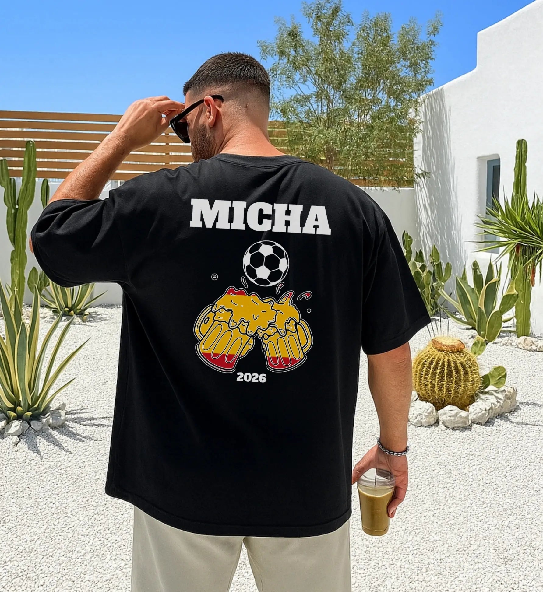 Personalisiertes schwarzes Herren T-Shirt mit Aufdruck 'MICHA', einem Fußball und zwei anstoßenden Bierkrügen in den spanischen Nationalfarben vor einer mediterranen Kulisse.