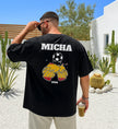 Personalisiertes schwarzes Herren T-Shirt mit Aufdruck 'MICHA', einem Fußball und zwei anstoßenden Bierkrügen in den spanischen Nationalfarben vor einer mediterranen Kulisse.