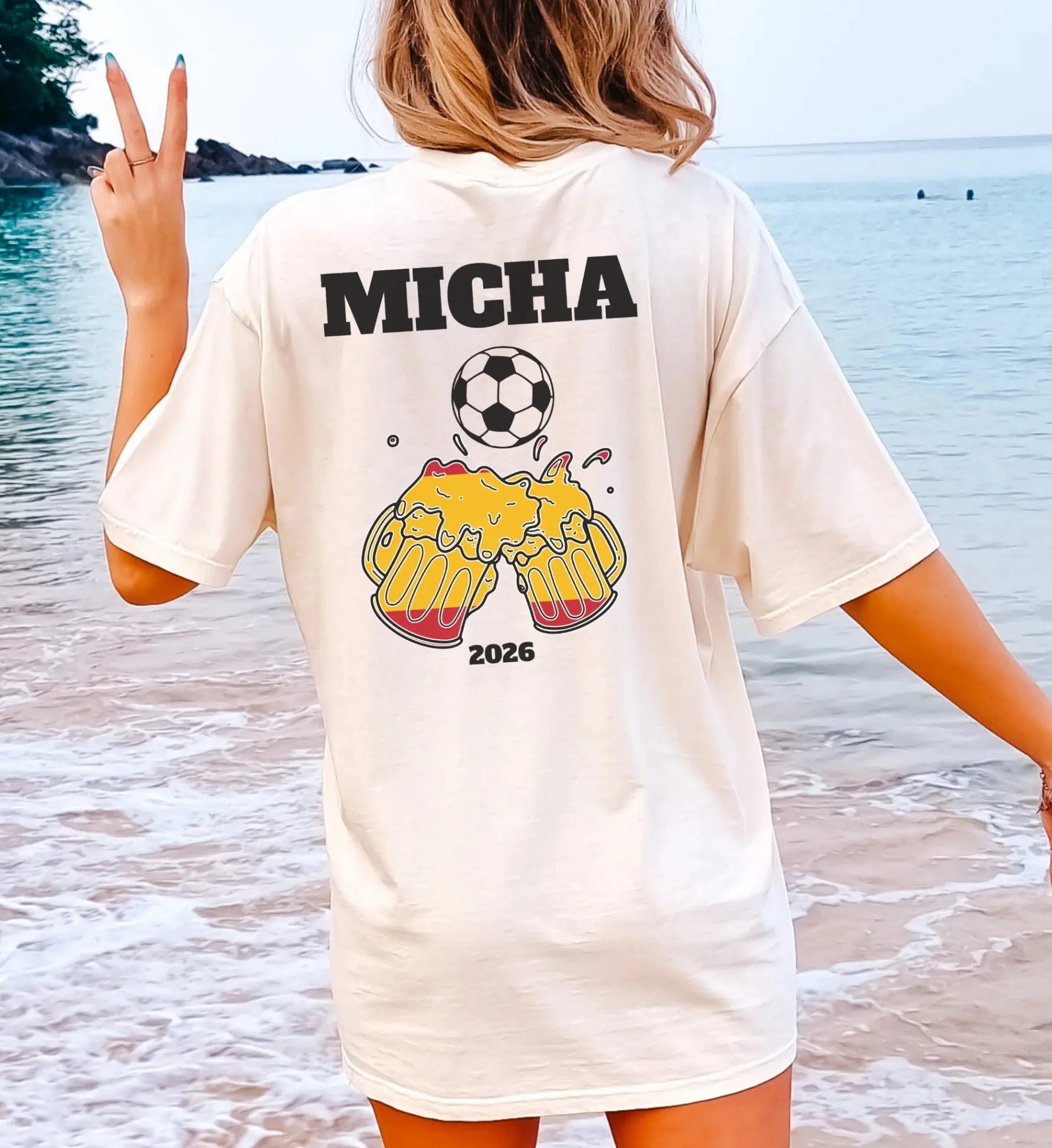 Frau am Strand in einem personalisierten cremefarbenen Oversize T-Shirt mit dem Namen Micha, einem Fußball und anstoßenden Bierkrügen im Spanien-Design für 2026.
