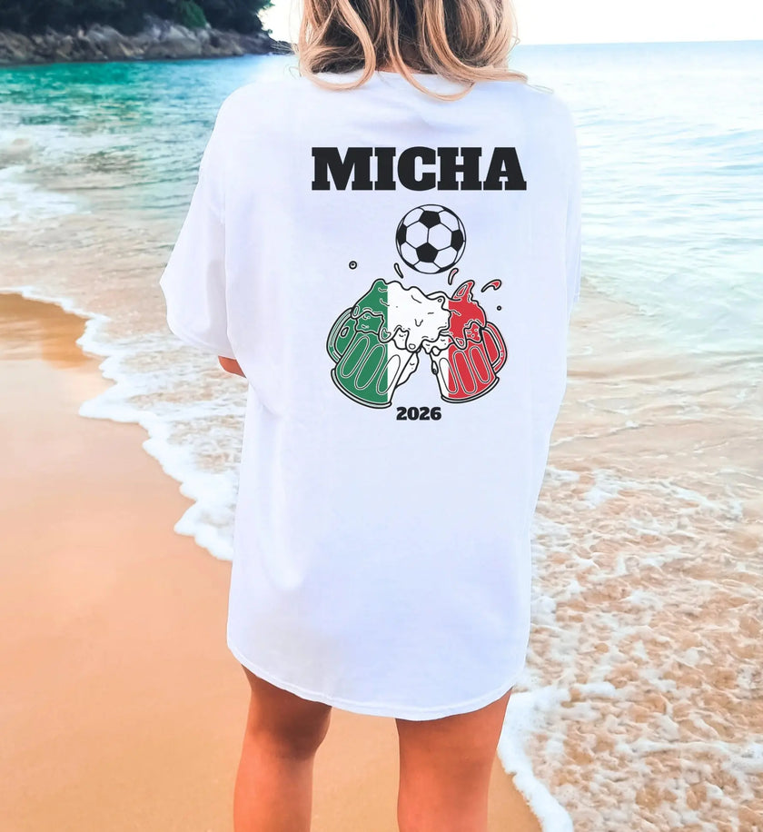 Weißes personalisiertes T-Shirt mit Rückenprint für Italien-Fans, Motiv mit Fußball, anstoßenden Bierkrügen in den italienischen Landesfarben, dem Namen Micha und der Jahreszahl 2026 vor einer Strandkulisse.