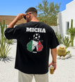 Ein Mann trägt ein schwarzes, personalisiertes T-Shirt mit der Aufschrift 'MICHA', einem Fußball und zwei anstoßenden Bierkrügen in den Nationalfarben Italiens (Grün, Weiß, Rot) sowie der Jahreszahl 2026.