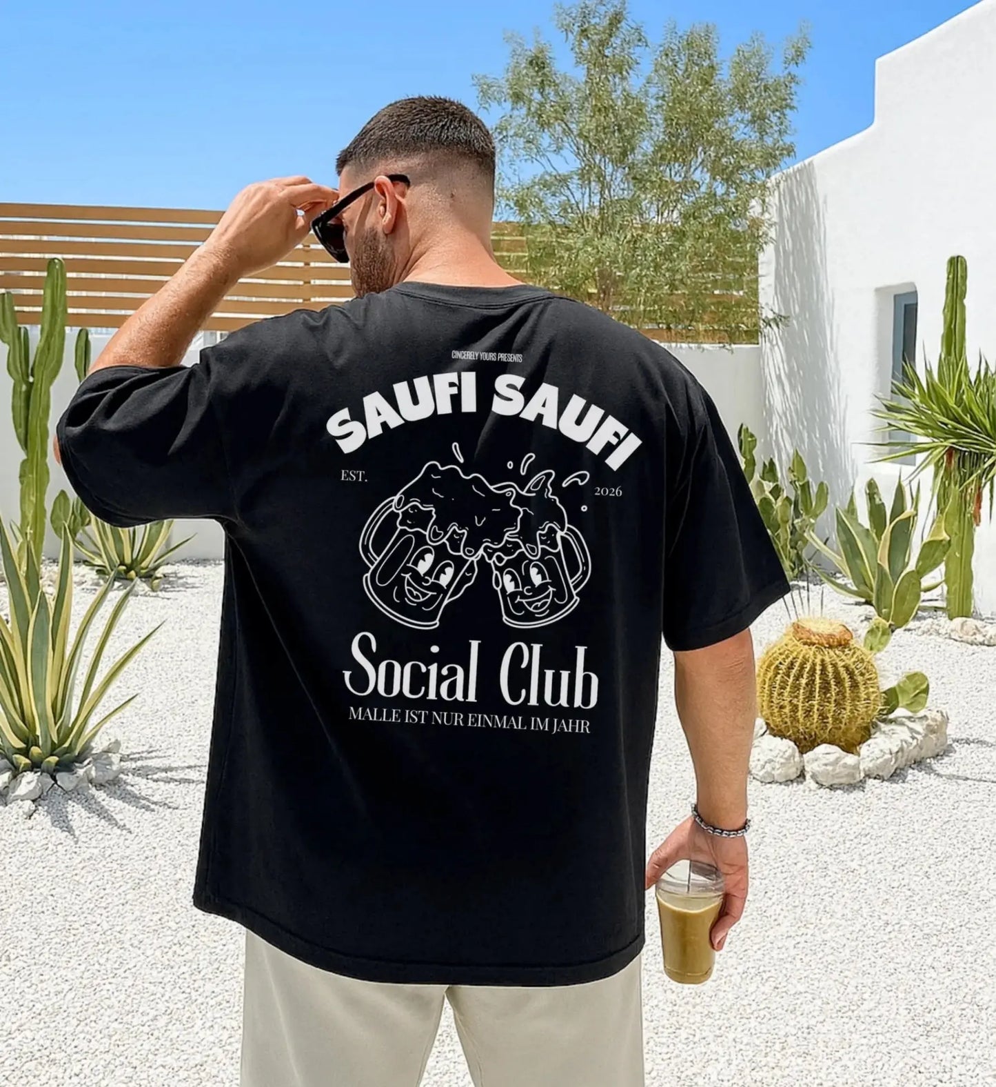 Ein Mann von hinten in einem schwarzen Oversized T-Shirt mit dem weißen Aufdruck 'Saufi Saufi Social Club', lachenden Bierkrügen und dem Schriftzug 'Malle ist nur einmal im Jahr' in einem sommerlichen Garten mit Kakteen.