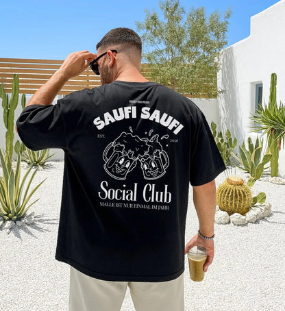 Ein Mann von hinten in einem schwarzen Oversized T-Shirt mit dem weißen Aufdruck 'Saufi Saufi Social Club', lachenden Bierkrügen und dem Schriftzug 'Malle ist nur einmal im Jahr' in einem sommerlichen Garten mit Kakteen.