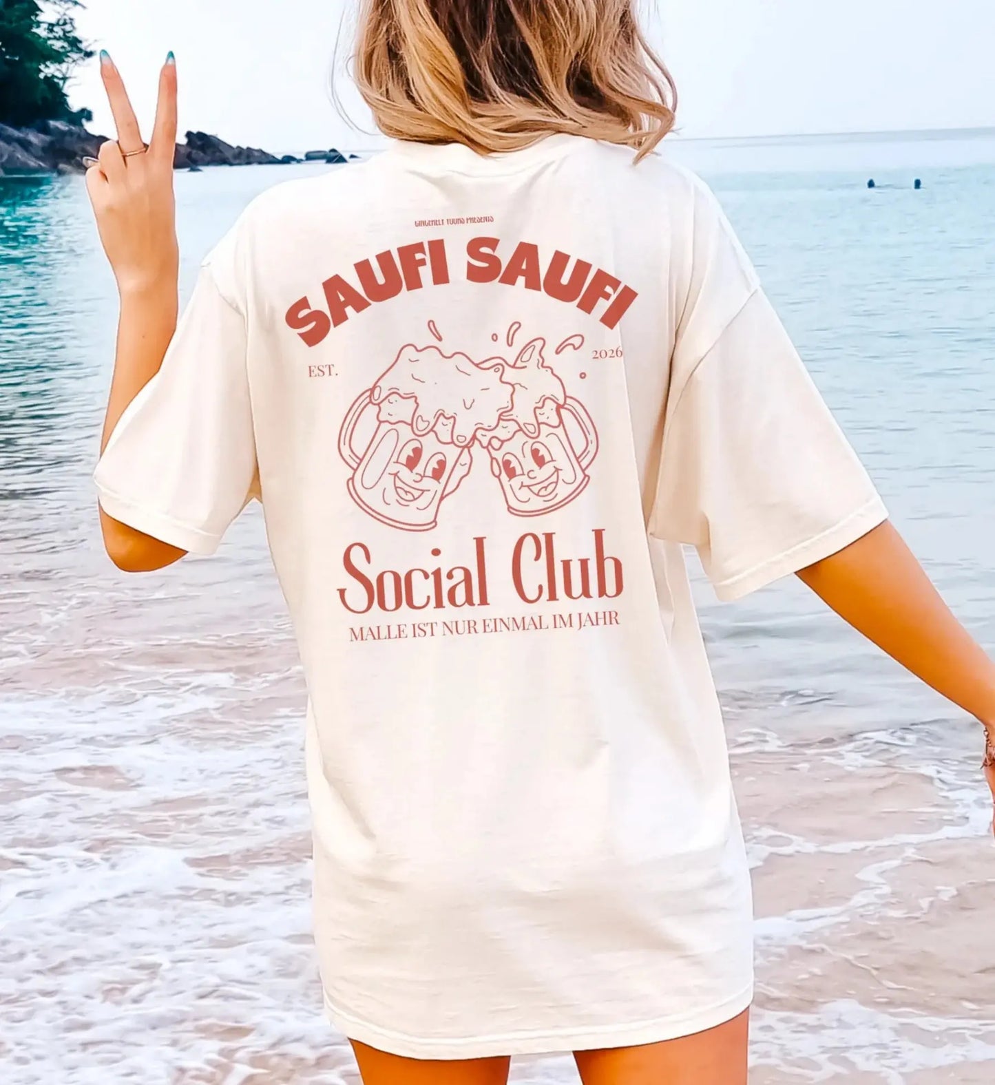 Rückansicht eines cremefarbenen Oversized T-Shirts mit Retro-Grafik zweier lachender Bierkrüge und dem Schriftzug Saufi Saufi Social Club am Strand.