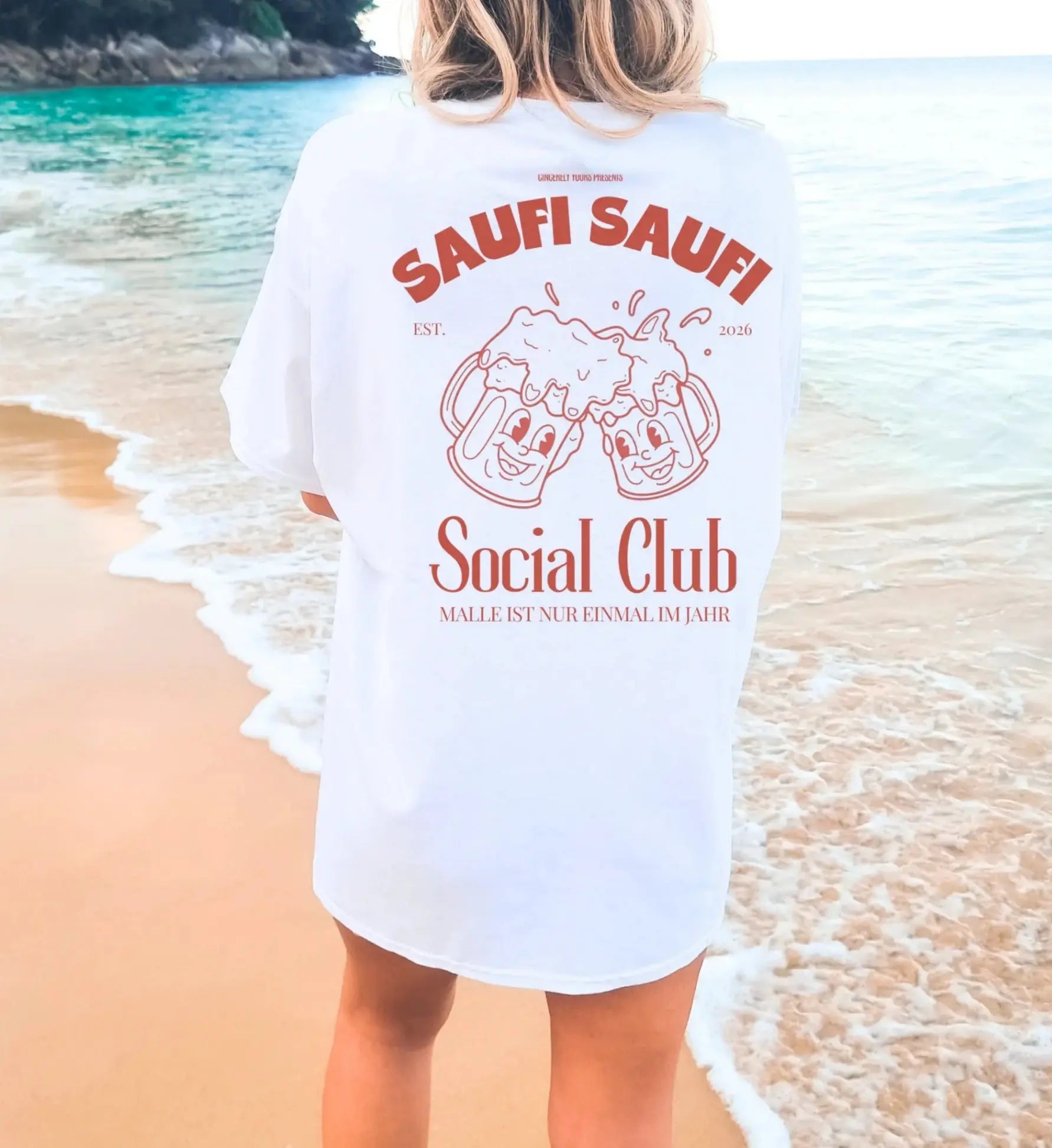Weißes Oversized T-Shirt mit rostbraunem Saufi Saufi Social Club Rückenprint und lachenden Bierkrug-Maskottchen am Strand.