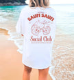 Weißes Oversized T-Shirt mit rostbraunem Saufi Saufi Social Club Rückenprint und lachenden Bierkrug-Maskottchen am Strand.